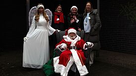 Nikolaus sitzt im Sessel, Engel und QM-Teammitglieder stehen daneben auf dem Wintermarkt in der Thermometersiedlung.