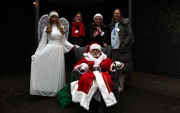 Nikolaus sitzt im Sessel, Engel und QM-Teammitglieder stehen daneben auf dem Wintermarkt in der Thermometersiedlung.