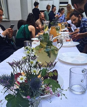 Eine Gruppe von Menschen sitzt an einem langen Tisch mit Getränken, Blumen und Speisen bei einem Nachbarschaftsdinner im Hinterhof.