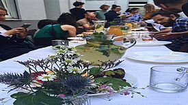 Eine Gruppe von Menschen sitzt an einem langen Tisch mit Getränken, Blumen und Speisen bei einem Nachbarschaftsdinner im Hinterhof.