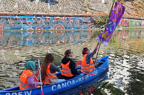 Vier Personen in orangefarbenen Westen sitzen in einem blauen Kanu mit bunter Fahne in einem Kanal.