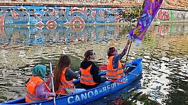 Vier Personen in orangefarbenen Westen sitzen in einem blauen Kanu mit bunter Fahne in einem Kanal.