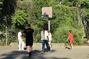 Fünf junge Menschen spielen draußen Basketball. Rückenansicht vor dem Basketballkorb. Im Hintergrund Bäume und Sträucher. Ein Spieler im Vordergrund trägt ein T-Shirt mit der Aufschrift „Team“.