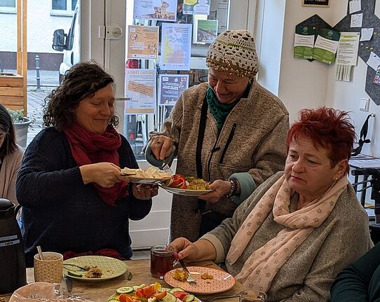 Zwei Frauen sitzen beim internationalen Frühstück am Tisch während Sedanur Karaca neben ihnen steht und sich etwas zu Essen nimmt.