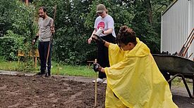 Drei Personen arbeiten an einem Beet im Freien, eine Person steckt Markierungen in den Boden.