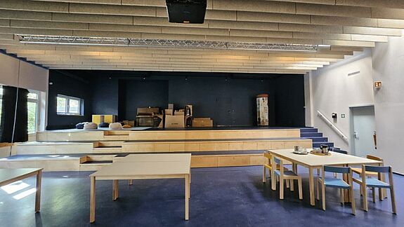 In einem hellen Saal stehen Tische und Stühle vor einer Bühne mit Sitzstufen und dunkler Rückwand.