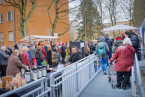 Ca. 40 Menschen unterschiedlichen Alters meist vom hinten sichtbar. Sie laufen vorbei an einem überdachten Stand mit Wimpeln, an einem Kuchen- und Kaffeebuffet, einige mit Rollatoren auf einer Metallrampe an einem blauen Containerbau. Im Hintergrund Bäume und mehrstöckige Wohnneubauten. 