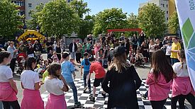 Viele Kinder und Erwachsene vergnügen sich bei sonnigem Wetter auf einem Platz, der von Bäumen und Wohngebäuden gesäumt ist.  