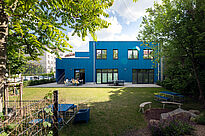 Im Vordergrund liegen eine Rasenfläche, Beete und mehrere Sitzgelegenheiten. Dahinter steht das Blueberry-Gebäude mit blauer Fassade, vor dem einige Menschen an einem Tisch auf der gepflasterten Terrasse sitzen.
