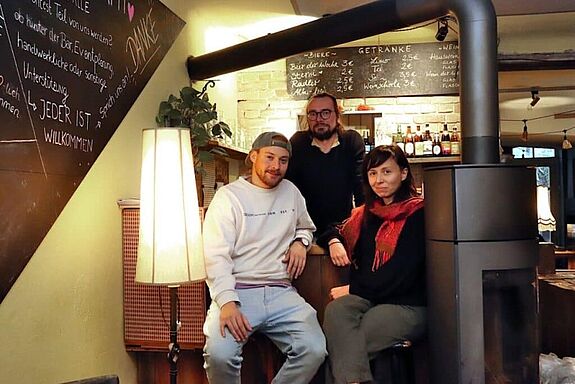 Zwei Engagierte sitzen an der Bar im Kieztreff „Kamine&Wein“, eine weitere Person steht hinter ihnen. 