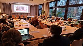 Menschen sitzen bei einer öffentlichen Quartiersratssitzung an einem großen U-förmigen Tisch und folgen einer Präsentation.