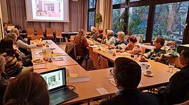 Menschen sitzen bei einer öffentlichen Quartiersratssitzung an einem großen U-förmigen Tisch und folgen einer Präsentation.
