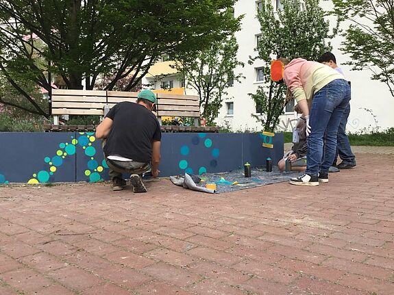 Kinder und Erwachsene bemalen gemeinsam mit Sprühdosen eine Betonbank am Satellitenplatz mit bunten Kreisen und Mustern.