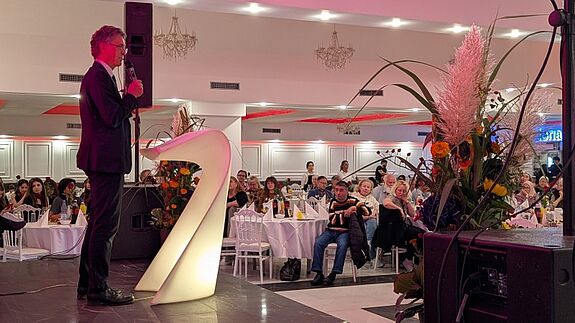 Ein Redner im Anzug spricht am Mikrofon auf einer Bühne vor einem großen Saal mit Publikum; neben ihm steht eine beleuchtete Zahl „2“ und ein Blumengesteck.