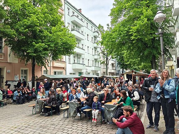 Viele Menschen sitzen und stehen an Tischen zwischen Marktständen auf einer gesperrten, von Bäumen gesäumten Straße.