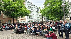 Viele Menschen sitzen und stehen an Tischen zwischen Marktständen auf einer gesperrten, von Bäumen gesäumten Straße.