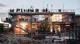 Die Außenansicht eines Gebäudes mit großen Glasflächen am Abend, auf dessen Dach, Terrassen und Außentreppe Menschen stehen. Im Vordergrund abgeschirmt durch einen Zaun liegen Baustoffe. 