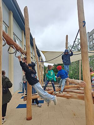 Eine Holzkonstruktion im Außenraum mit Kletterseilen, Balancierbalken und Sonnensegel. Sechs Kinder spielen darauf. Am linken Rand stehen zwei Erwachsene. Im Hintergrund Bäume und Wohnungsneubau, links im Bild die Außenwand eines Funktionsbaus mit Fenstern.  