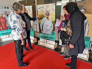 Vier weibliche Personen auf einem roten Teppich stehend schauen sich gemeinsam eine Ausstellung an und sprechen miteinander. Auf den ausgestellten stehenden Tafeln sind Portraits von Menschen abgebildet. Eine Person zeigt auf ein Portrait einer älteren Frau, die lächelt. Unter ihrem Foto steht „Willkommen hier, wo Vielfalt wohnt“.   