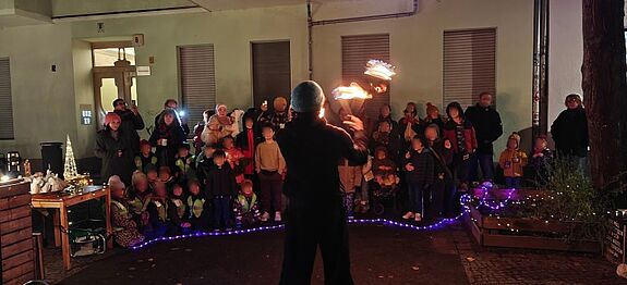 Feuerkünstler jongliert brennende Stäbe, während Kinder und Erwachsene im Halbkreis vor dem Quartiersbüro zuschauen.