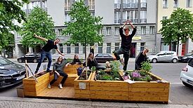 Mehrere Personen sitzen und posieren auf einem bepflanzten Holzparklet im Pankkiez.