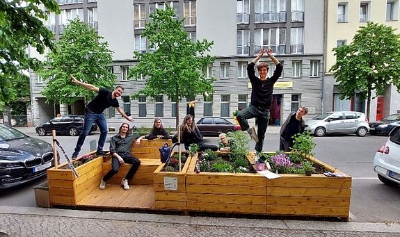 Mehrere Personen sitzen und posieren auf einem bepflanzten Holzparklet im Pankkiez.