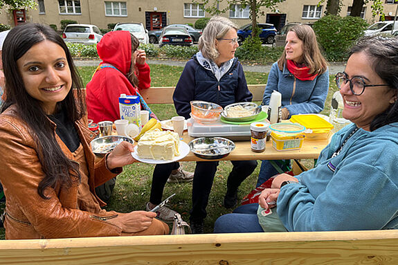 Zwei Frauen auf einer Sitzbank beim Frühstücken. Im Hintergrund drei weitere Frauen, die sich unterhalten. 