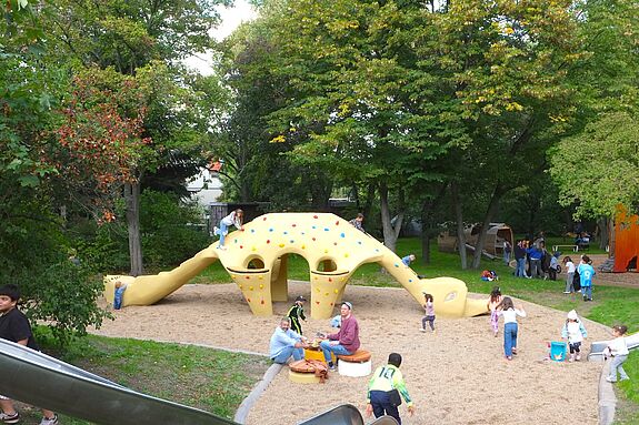 Ein Spielplatz umringt von Bäumen mit einem gelben Dinosaurier als Klettergerüst. Im Vordergrund ist eine Rutsche. Kinder spielen auf der Fläche. 