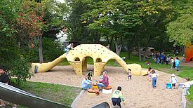 Ein Spielplatz umringt von Bäumen mit einem gelben Dinosaurier als Klettergerüst. Im Vordergrund ist eine Rutsche. Kinder spielen auf der Fläche. 
