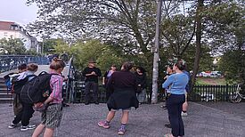 Eine Gruppe von etwa zehn Menschen steht auf einem Gehweg neben einer Brücke und hört einer Person zu, die etwas erklärt. 