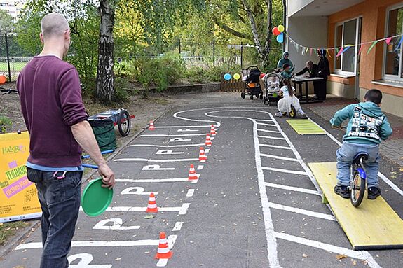 Ein Kind fährt mit dem Fahrrad über eine gelbe Wippe, die Teil eines Fahrradparcours ist, während ein Mann zuschaut.