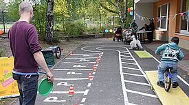 Ein Kind fährt mit dem Fahrrad über eine gelbe Wippe, die Teil eines Fahrradparcours ist, während ein Mann zuschaut.