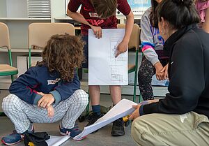 Drei Kinder und eine erwachsene Person hockend, sitzend und stehend. Das stehende Kind hält ein A3-Blatt mit einer Zeichnung vor dem Körper und zeigt darauf. Die anderen schauen auf das Blatt. Gesichter sind nicht erkennbar. Im Hintergrund Stühle