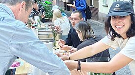 Mehrere Menschen sitzen und stehen an einer langen gedeckten Tafel im Freien und Essen gemeinsam.