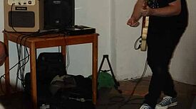 Ein Mann steht mit einer elektronischen Gitarre an einem Verstärker und Lautsprecher; Effektgeräte und ein kleines Keyboard liegen auf dem Boden.