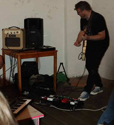 Ein Mann steht mit einer elektronischen Gitarre an einem Verstärker und Lautsprecher; Effektgeräte und ein kleines Keyboard liegen auf dem Boden.