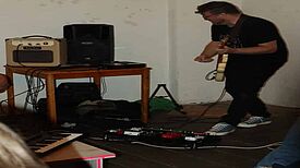 Ein Mann steht mit einer elektronischen Gitarre an einem Verstärker und Lautsprecher; Effektgeräte und ein kleines Keyboard liegen auf dem Boden.