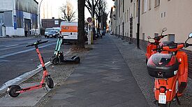 E-Roller und ein Mofa stehen auf einem Gehweg und stellen für die Menschen im Kiez ein Hindernis dar. 