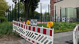 Bauzaun mit Absperrung und Warnlichtern steht vor einem eingezäunten Bolzplatz am Spielplatz Bendastraße.