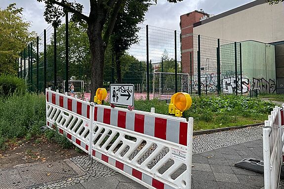 Bauzaun mit Absperrung und Warnlichtern steht vor einem eingezäunten Bolzplatz am Spielplatz Bendastraße.