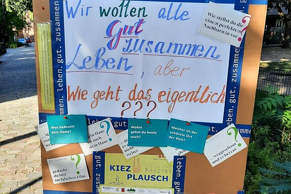 Auf einer Stellwand des Kiezplauschs hängt ein großes Plakat mit der Frage „Wir wollen alle gut zusammenleben, aber wie geht das eigentlich?“; daneben sind Karten mit Gesprächsfragen angeheftet.