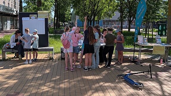 Eine Gruppe Jugendlicher steht an einem sonnigen Tag auf einem freien Platz vor Tischen mit dem Angebot „Back dir deinen Kiez“. 