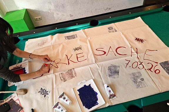 Eine Person bemalt ein großes Tuch auf einem Billardtisch mit dem Schriftzug „Starke Sache“.