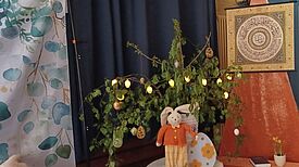 Ein Osterstrauch mit bunten Eiern und Lichterkette steht neben einem Stoffhasen und einer kleinen Blumendekoration.