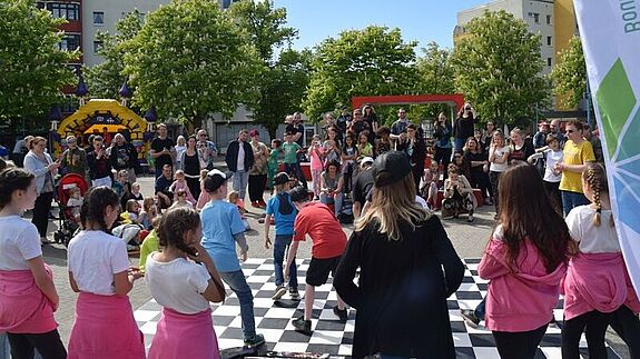 Viele Kinder und Erwachsene vergnügen sich bei sonnigem Wetter auf einem Platz, der von Bäumen und Wohngebäuden gesäumt ist.  
