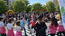 Viele Kinder und Erwachsene vergnügen sich bei sonnigem Wetter auf einem Platz, der von Bäumen und Wohngebäuden gesäumt ist.  