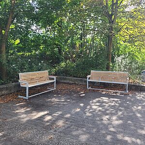 Zwei Bänke aus Holz und Metall stehen im rechten Winkel zueinander unter Baumen. Die Bänke haben jeweils eine Rückenlehne und mehrere Armlehnen.