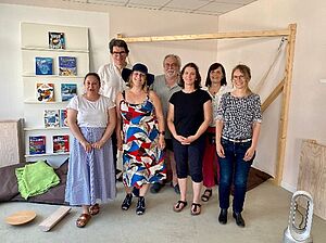 Gruppenfoto von sieben Menschen unterschiedlichen Alters in einem hellen Raum. Alle lächeln in die Kamera. Im Hintergrund erkennbar ist ein Regal mit Broschüren. 