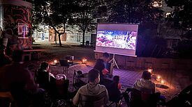 Menschen sitzen abends im Freien vor einer Leinwand, umgeben von Kerzenlicht, und schauen beim Freiluftkino den Film „Coco“.