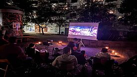 Menschen sitzen abends im Freien vor einer Leinwand, umgeben von Kerzenlicht, und schauen beim Freiluftkino den Film „Coco“.